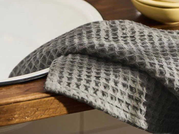 Et gråt håndklæde med vaffelstruktur hænger på kanten af en træbordplade nær en hvid vask.