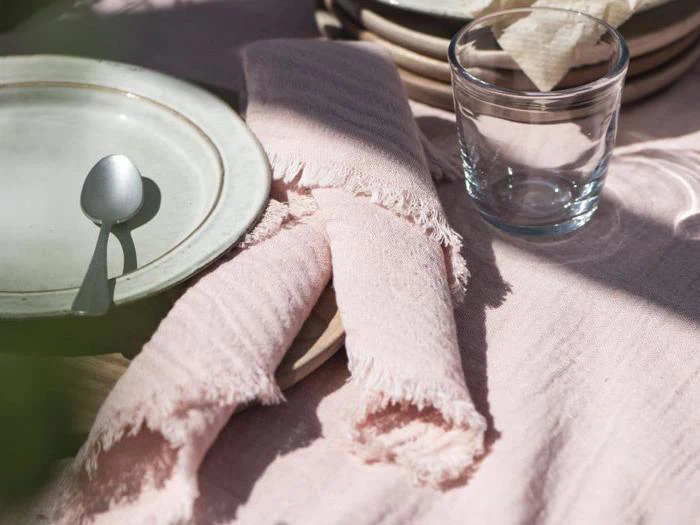 Nærbillede af et bord dækket med en hvid tallerken, en ske, et klart glas og en lyserød stofserviet på en matchende lyserød dug. Sollyset kaster bløde skygger over scenen.