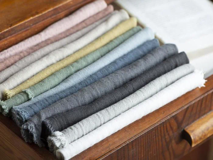 Pænt foldede hørstoffer i forskellige dæmpede farver er stablet i en træskuffe ved siden af en åben bog.