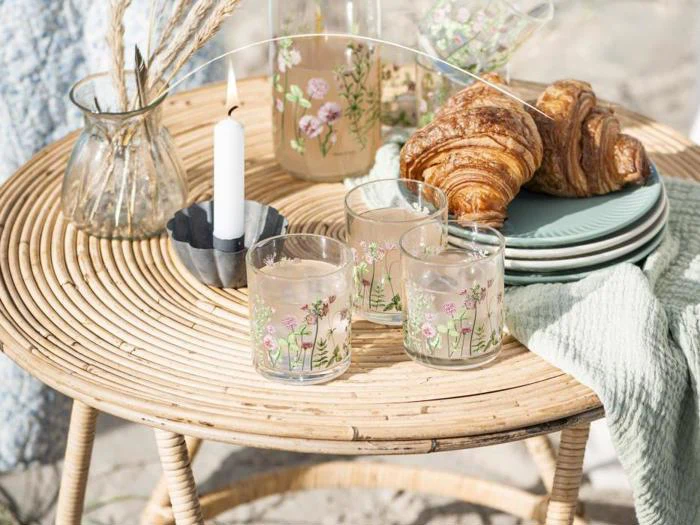 På et rundt kurvebord står blomsterglas med juice, en vase med tørret græs, et hvidt stearinlys, croissanter på grønne tallerkener og en sammenfoldet grøn dug i sollys.