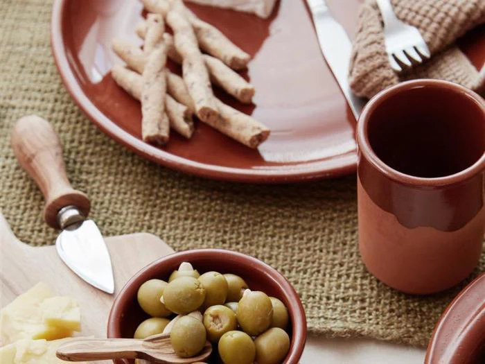 Nærbillede af en rustik borddækning med grønne oliven i en lille skål, brødstænger, en kop, ost, en ostekniv og en gaffel på brunt og beige service.