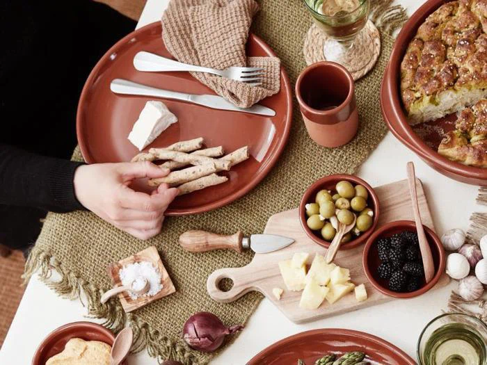 En person rækker ud efter brødstænger på et rustikt bord dækket med ost, oliven, bær og brød på brune keramikfade og en træplade.