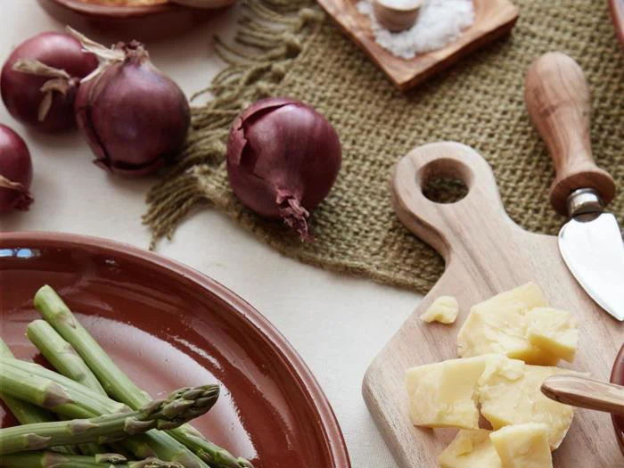 Hakket ost på et træbræt med en kniv, friske asparges på en brun tallerken og hele rødløg på et bord med en vævet måtte.