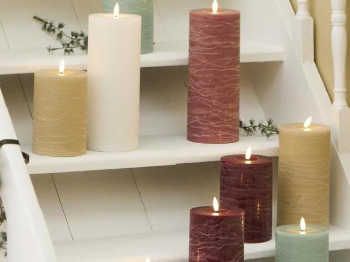 Flere store, tændte bloklys i forskellige farver er anbragt på en hvid trætrappe med nogle grønne kviste til pynt.