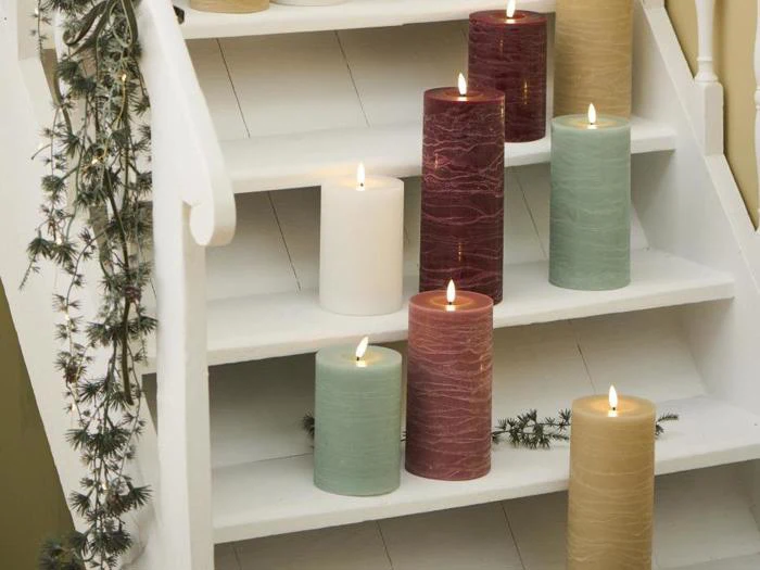 Flere store, tændte bloklys i rød, grøn, hvid og beige er anbragt på en hvid trætrappe, der er dekoreret med grønt.