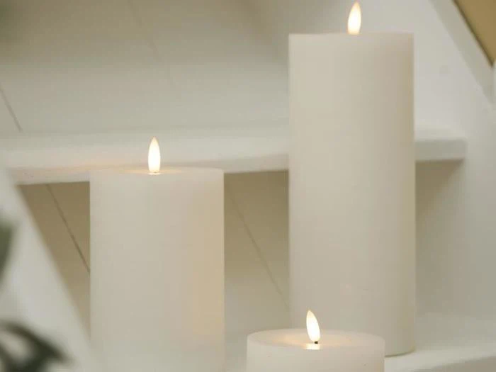 Tre hvide bloklys med flammer er placeret på hvide trin og skaber en blød og fredfyldt stemning.