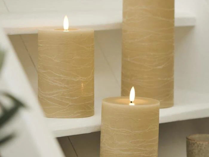 Tre tændte beige bloklys med strukturerede overflader er placeret på hvide trin og skaber en varm og hyggelig stemning.