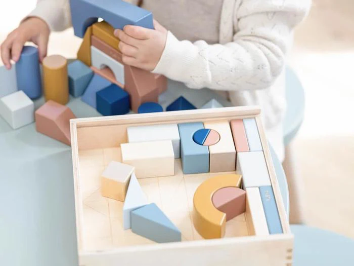 Et barn leger med pastelfarvede træbyggeklodser, som det anbringer i en træbakke på et lyseblåt bord.