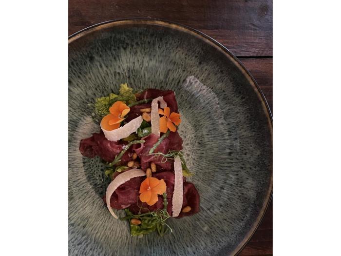 Tynde skiver råt kød garneret med orange spiselige blomster, grønt, pinjekerner og høvlet ost på en struktureret grøn tallerken.