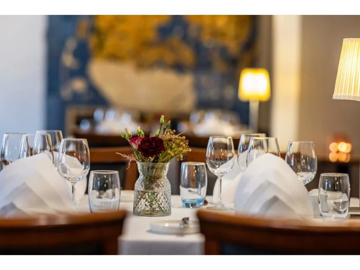 Elegant restaurantbord dækket med vinglas, vandglas, hvide servietter og en vase med røde blomster, med blød belysning og sløret baggrund.