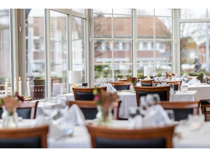 Lys, elegant restaurant med store vinduer, opdækkede borde med hvide duge, glas og blomsterdekorationer med udsigt over en gade udenfor.