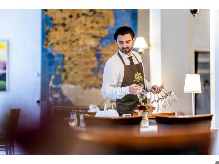 En tjener i hvid skjorte og mørkt forklæde dækker bord i en veloplyst restaurant med flere glas i hånden.
