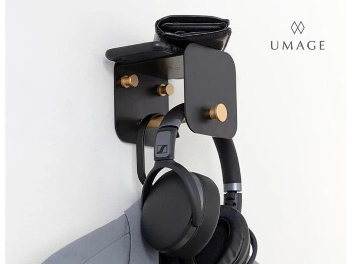 En vægknage i sort metal med gulddetaljer holder pung, telefon, hovedtelefoner og en hængende jakke. UMAGE-logoet er synligt i øverste højre hjørne.