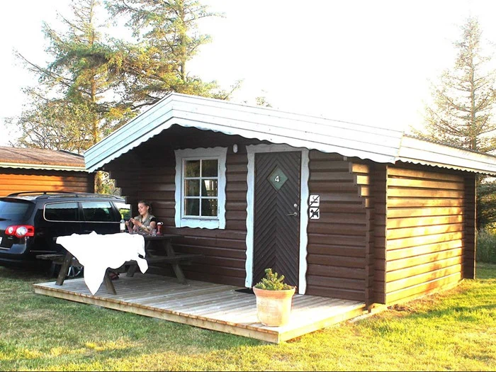 En person sidder ved et picnicbord foran en lille træhytte med en potteplante ved døren og en sort bil parkeret i nærheden, omgivet af træer og græs.