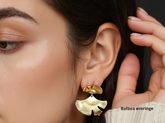 Et nærbillede af en kvinde med en guldvifteformet ørering, mens hendes hånd rører ved hendes øre. Teksten på billedet lyder Balboa øreringe.