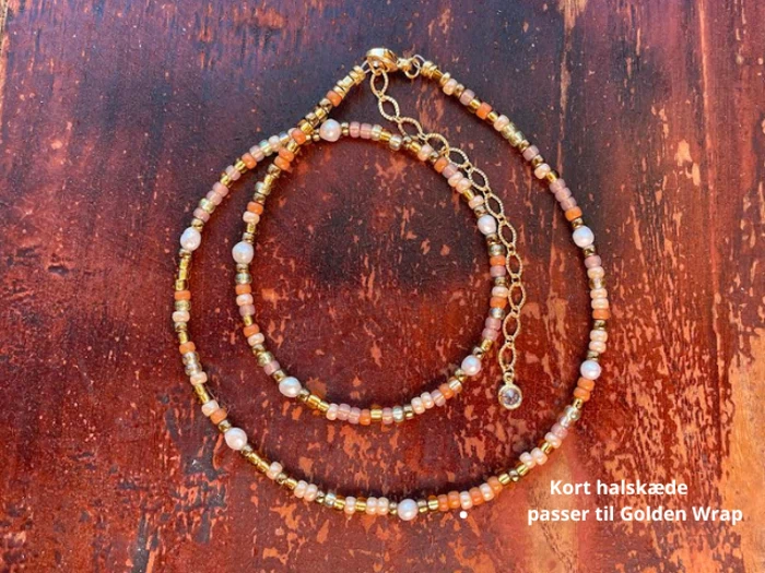 En kort halskæde med guld-, brun- og perlelignende perler er lagt på en træoverflade. Den danske tekst lyder: "Kort halskæde passer til Golden Wrap.".