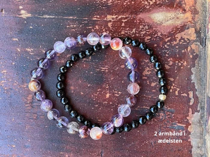 To perlearmbånd af ædelsten på en rustik træoverflade; det ene armbånd har lilla/klare perler, det andet har små sorte perler. Teksten på dansk lyder 2 armbånd i ædelsten.