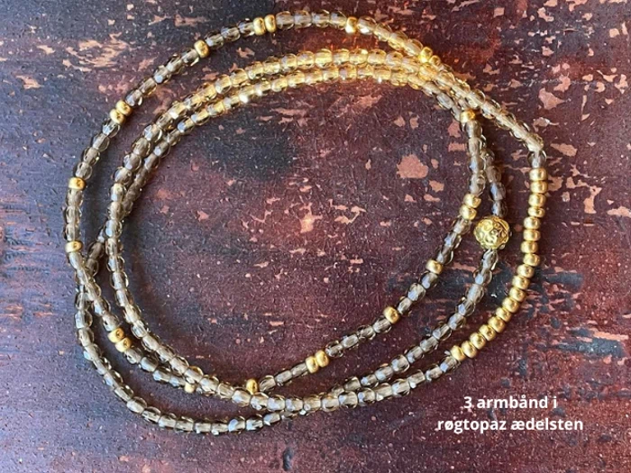 Tre armbånd af røgtopas-ædelsten og små guldfarvede perler er udstillet på en rustik, brun træoverflade. Den danske tekst i hjørnet lyder: 3 armbånd i røgtopaz ædelsten.