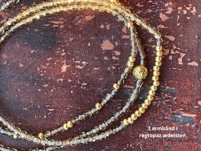 Nærbillede af tre armbånd lavet af røgtopas og guldfarvede perler på en struktureret brun overflade. Teksten lyder: 3 armbånd i røgtopaz ædelsten.