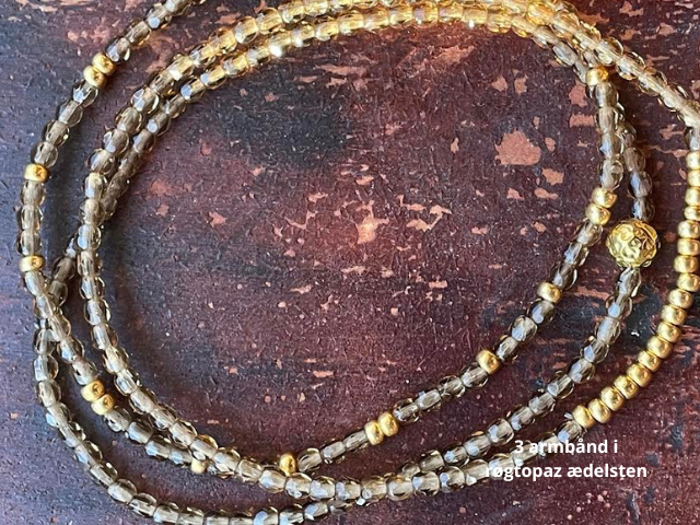 Tre perlearmbånd i lys topas ædelsten og guldaccenter, oprullet på en mørk, struktureret overflade. Dansk tekst i hjørnet lyder: 3 armbånd i topaz ædelsten.