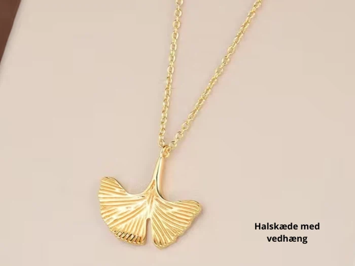 En guldhalskæde med et vedhæng formet som et ginkgoblad, vist på en beige overflade. Teksten på dansk lyder: Halskæde med vedhæng.