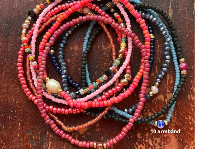 Flere farverige perlearmbånd, de fleste i røde, rosa og mørkeblå nuancer, er stablet i en bunke på en brun træoverflade. Teksten lyder 10 armbånd.
