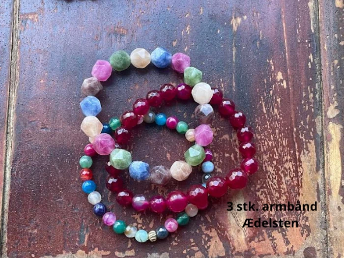 Tre farverige perlearmbånd af ædelsten er udstillet på en rustik træoverflade. Den danske tekst lyder: 3 stk. armbånd Ædelsten.