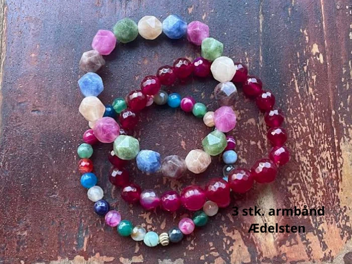 Tre perlearmbånd af ædelsten i forskellige farver er placeret på en rustik træoverflade. Sort tekst lyder, 3 stk. armbånd Ædelsten.