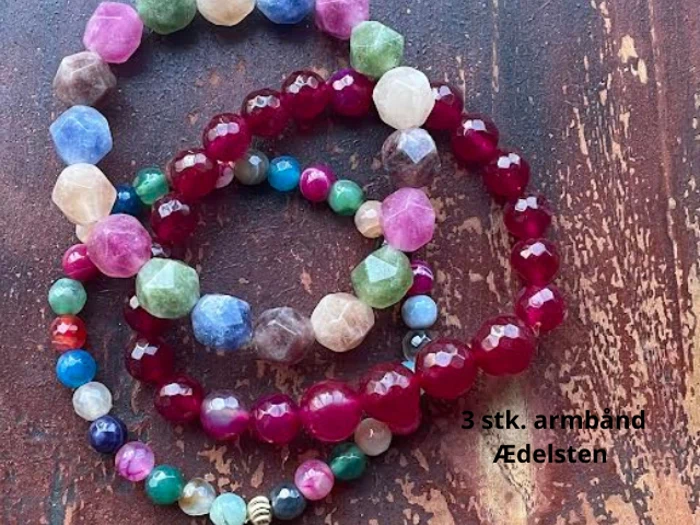 Tre farverige perlearmbånd med røde, pink, blå, grønne og hvide perler på en rustik brun overflade. Teksten lyder: 3 stk. armbånd Ædelsten.