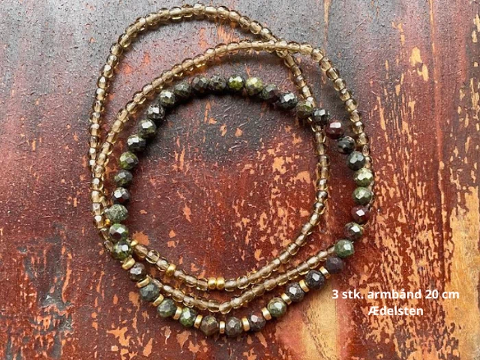 Tre perlearmbånd af ædelsten, hver 20 cm lange, er viklet på en mørk rødbrun træoverflade. Tekst på dansk er synlig i nederste højre hjørne.