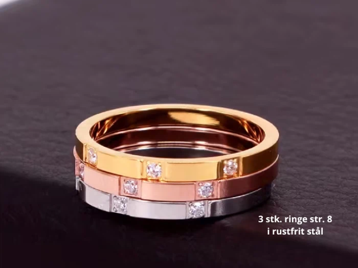 Tre ringe i rustfrit stål i guld-, rosaguld- og sølvfarver, hver med små indlejrede ædelsten, stablet på en mørk læderoverflade. Tekst på dansk er synlig i nederste højre hjørne.