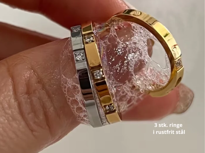 En hånd holder tre ringe i rustfrit stål - to i sølv og en i guld - alle med indlejrede sten og dækket af sæbebobler. Teksten lyder: 3 stk. ringe i rustfrit stål.