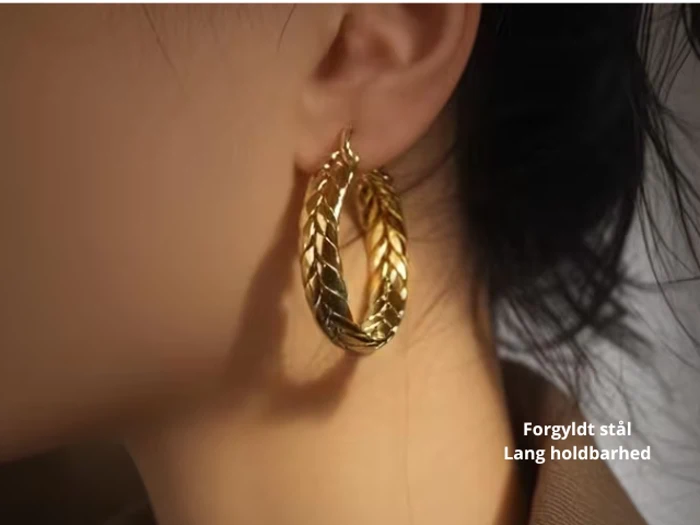 Nærbillede af en person iført store, guldfarvede, bladmønstrede øreringe. Teksten på dansk lyder: Forgyldt stål, Lang holdbarhed.