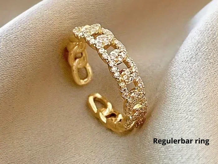 En guldring med åbent bånd og små klare krystaller på et silkeagtigt, beige stof. Ordene Regulerbar ring er trykt i hjørnet.