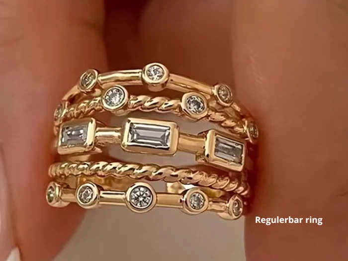 En guldring med flere bånd, runde og rektangulære klare ædelstene og snoede rebdetaljer, der holdes mellem to fingre. Teksten Regulerbar ring vises i hjørnet.