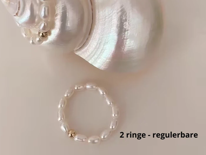 En fin perlering med en guldperle vises på en cremefarvet overflade ved siden af en hvid muslingeskal. Teksten 2 ringe - regulerbare står i nederste højre hjørne.