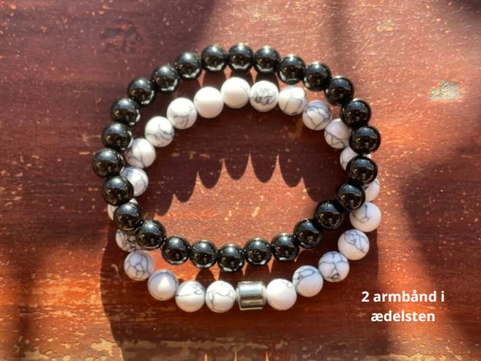 To perlearmbånd af ædelsten - et med sorte perler og et med hvide, marmorerede perler - på en træoverflade med sollys og skygge. Tekst: 2 armbånd i ædelsten.