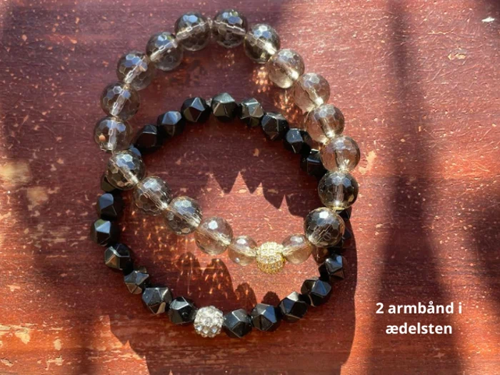 To perlearmbånd af ædelsten, et med klare runde perler og et med sorte facetterede perler, ligger på en træoverflade. Teksten på dansk lyder 2 armbånd i ædelsten.