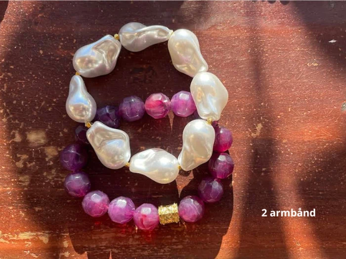 To armbånd på en træoverflade: et med store, uregelmæssige hvide perler og et med facetterede lilla perler og en guldaccent. Teksten lyder 2 armbånd.