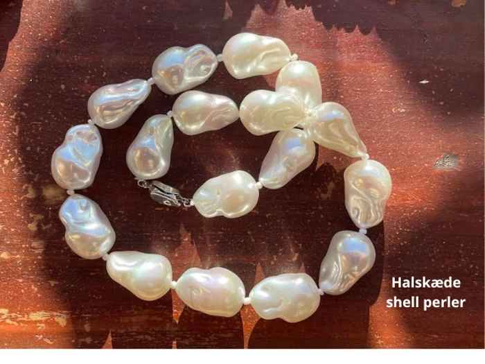 En halskæde af store, uregelmæssigt formede hvide skalperler vises på en træoverflade med sollys og skygger. Teksten lyder: Halskæde shell perler.