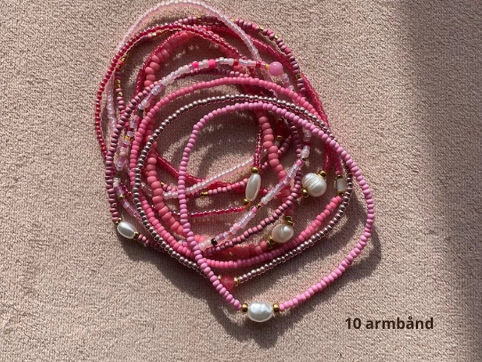 Ti lyserøde perlearmbånd, nogle med perler og gulddetaljer, er arrangeret i en bunke på en beige overflade. Teksten 10 armband står i nederste højre hjørne.