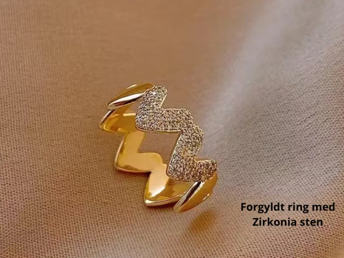 En guldbelagt ring med zigzag-design, prydet med zirkonia-sten, placeret på et beige stof. Teksten lyder: Forgyldt ring med Zirkonia sten.