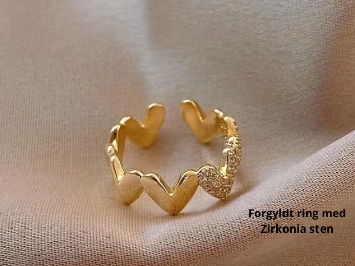 Guldbelagt ring med hjerteformede mønstre, det ene besat med små zirkonsten, placeret på et blødt beige stof. Tekst: Forgyldt ring med Zirkonia sten.