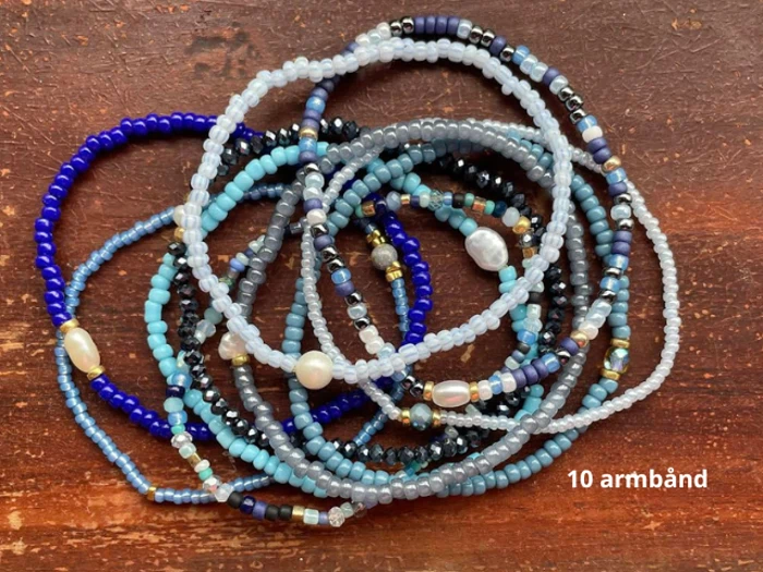 Ti perlearmbånd i blå, sorte, gyldne og hvide toner er arrangeret på en brun træoverflade. Teksten 10 armband står i nederste højre hjørne.