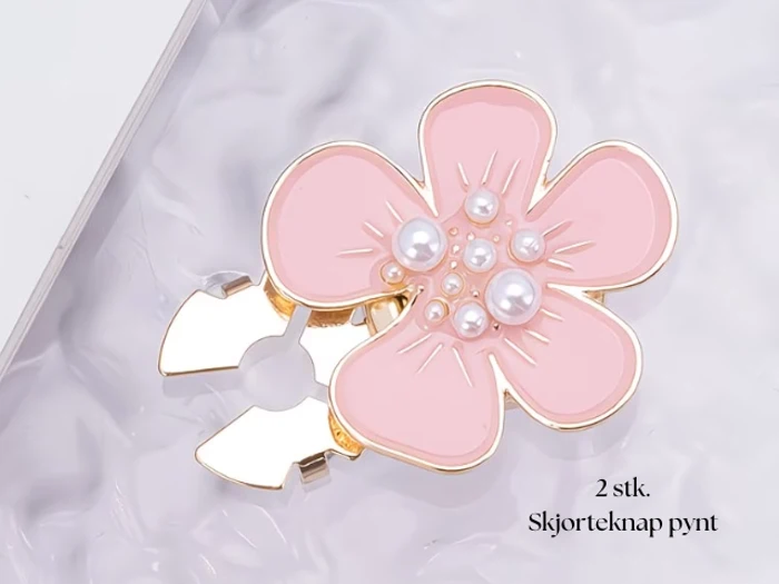 En dekorativ blomsterformet skjorteknap i guld og pink med perlemorsdetaljer vises på en struktureret overflade. Teksten lyder: 2 stk. Skjorteknap pynt.
