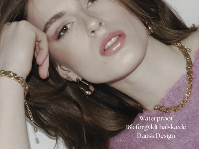 En kvinde med langt brunt hår bærer guldsmykker og en lilla top. Teksten lyder: Vandtæt 18k forgyldt halskæde Dansk Design.