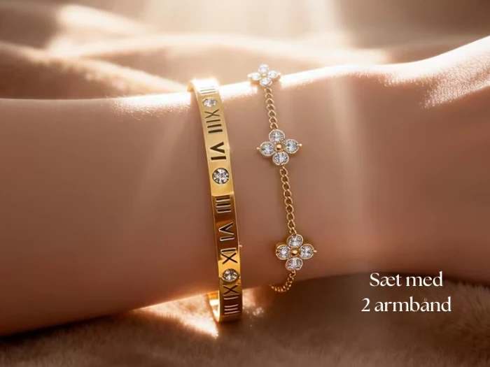 Et håndled med to guldarmbånd: et med romertal og et med små blomsterformede charms med krystaller. Teksten lyder: Sæt med 2 armbånd.