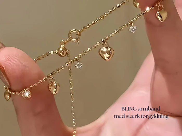 En hånd holder et fint guldkædearmbånd prydet med små hjertecharms og funklende ædelstene. Den danske tekst på billedet lyder: BLING armbånd med stærk forgyldning.