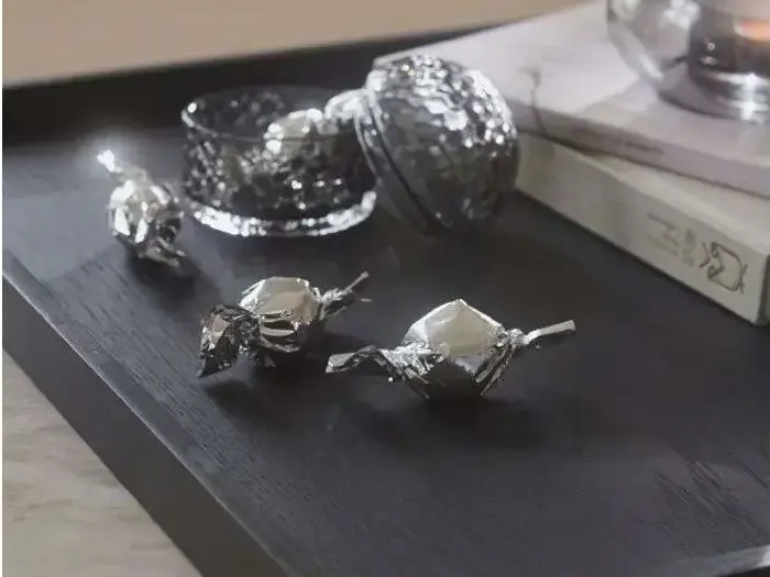 Tre sølvindpakkede bolsjer ligger på en sort bakke ved siden af en dekorativ glasbeholder og en bog med delvis synlig omslagstekst.