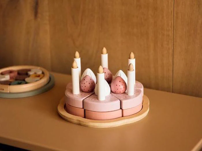 Fødselsdagskage af trælegetøj med lyserøde skiver, hvide lys og lyserøde jordbær, placeret på en træoverflade mod en brun baggrund.